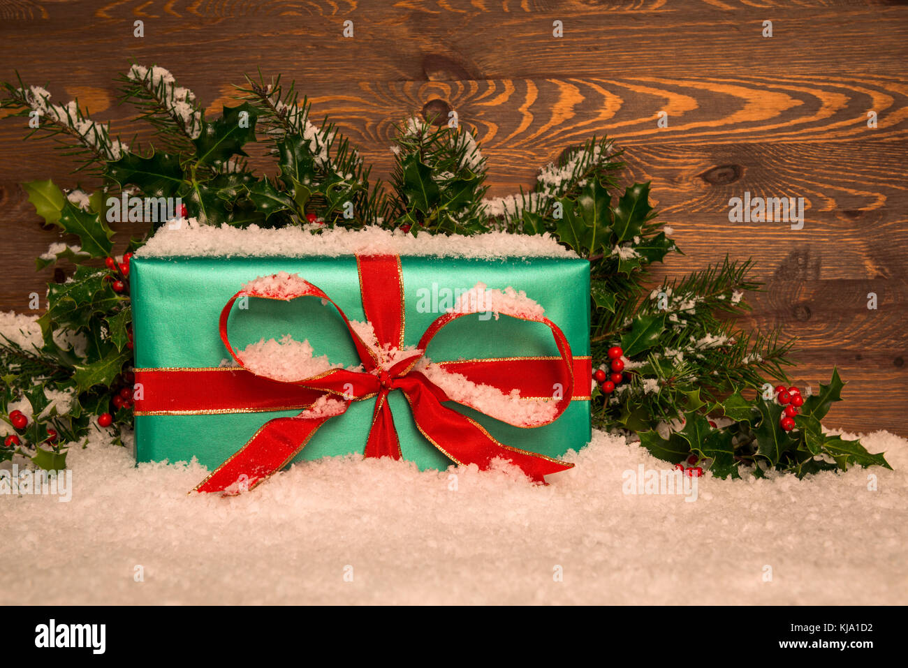 Weihnachtsgeschenk Geschenk im Grünbuch mit roter Schleife verpackt mit Stechpalme und Schnee gegen eine Holz- Hintergrund, kopieren Sie Platz für Ihre Nachricht. Stockfoto