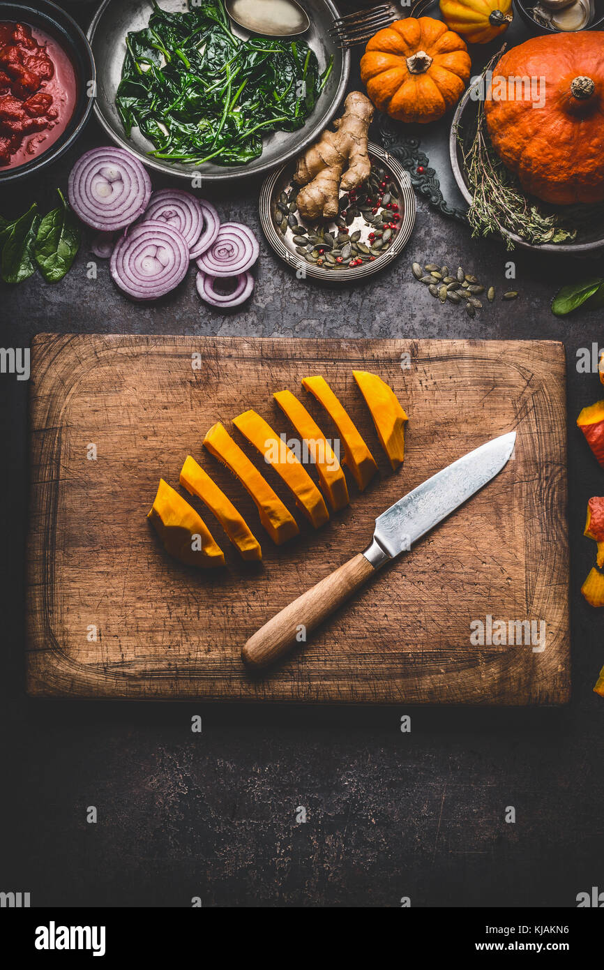Kürbis in Scheiben geschnitten auf Schneidebrett mit Messer und verschiedene Gemüse und Gewürze Zutaten für leckere saisonale Gericht kochen, rustikal, Ansicht von oben. Stockfoto