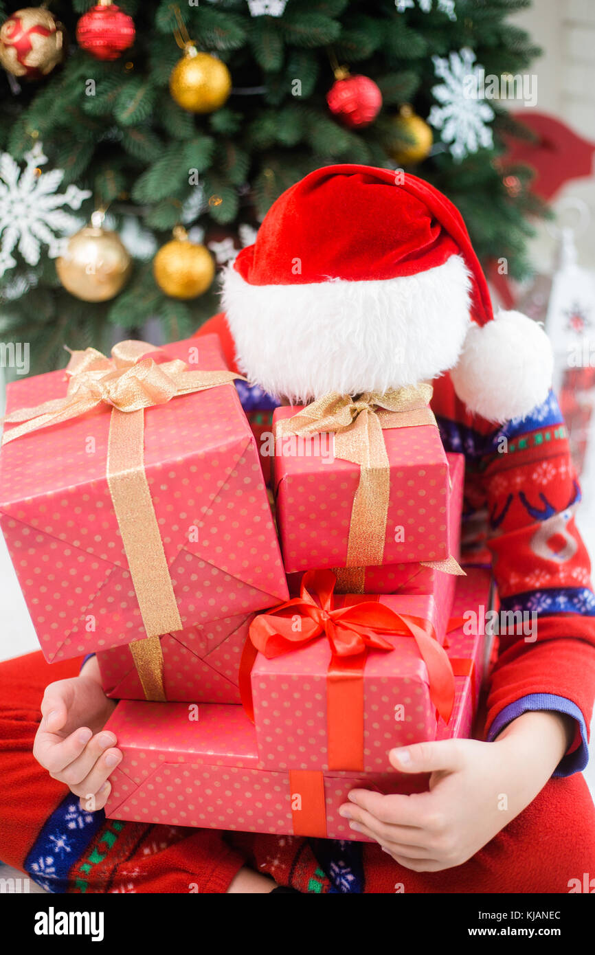 Lustige kleine Kind in Christmas Red hat sich hinter Haufen Urlaub präsentiert. Grün schöne Xmas Tree im Hintergrund. vertikale Farbfotografie. Stockfoto