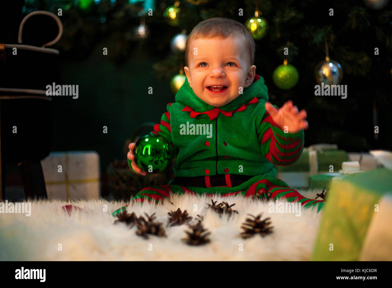 Happy little boy hält Christmas Ball in Hand, sitzt auf Fell Teppich und lächelt auf dem Hintergrund von Weihnachten Geschenkboxen und Feier. Stockfoto