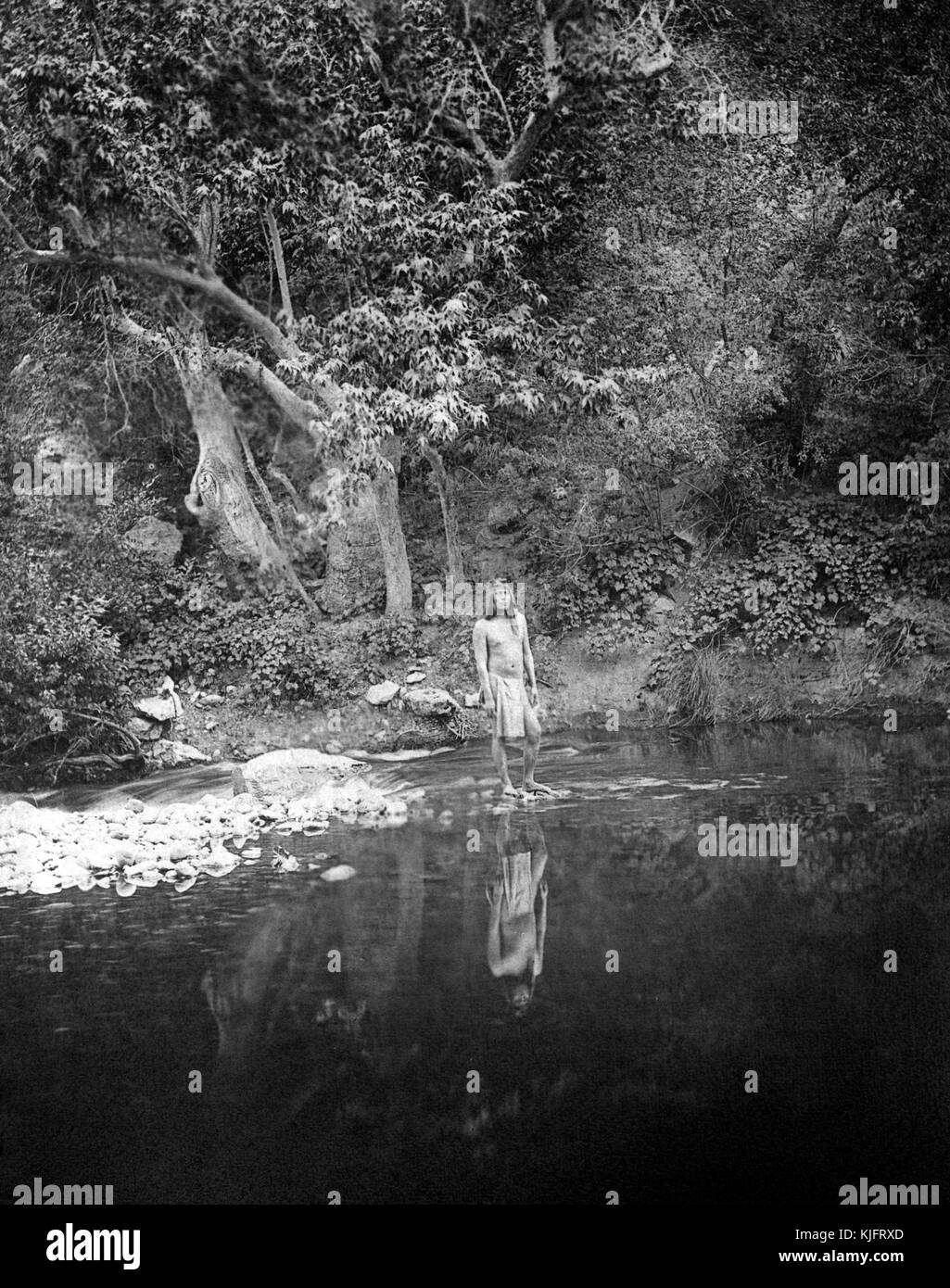 Ein Foto eines Mannes, der dem Stamm der Apache Indianer angehört, steht am Rande eines tiefen Wasserbeckens, seine Reflexion ist deutlich im Wasser zu sehen, Wasser fließt über Felsen hinter dem Mann, der Hintergrund besteht aus einem stark bewaldeten Gebiet, 1906. Aus der New York Public Library. Stockfoto