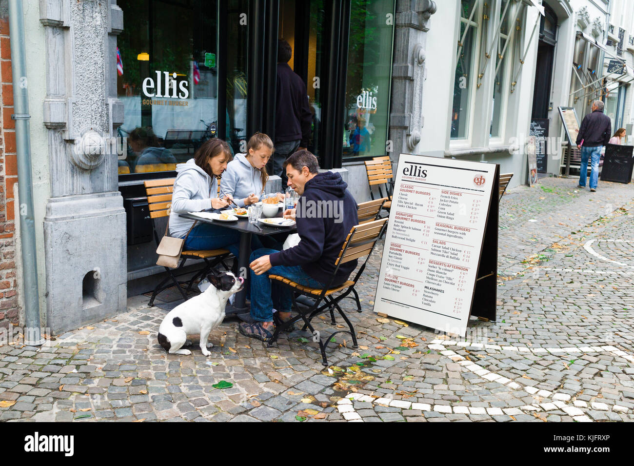 Eine Familie mit einem Hund außerhalb eines beliebten Burger Restaurant im Zentrum von Brügge, Flandern, Belgien essen. Der Hund am Tisch und seine Zunge heraus zugeführt wird Stockfoto