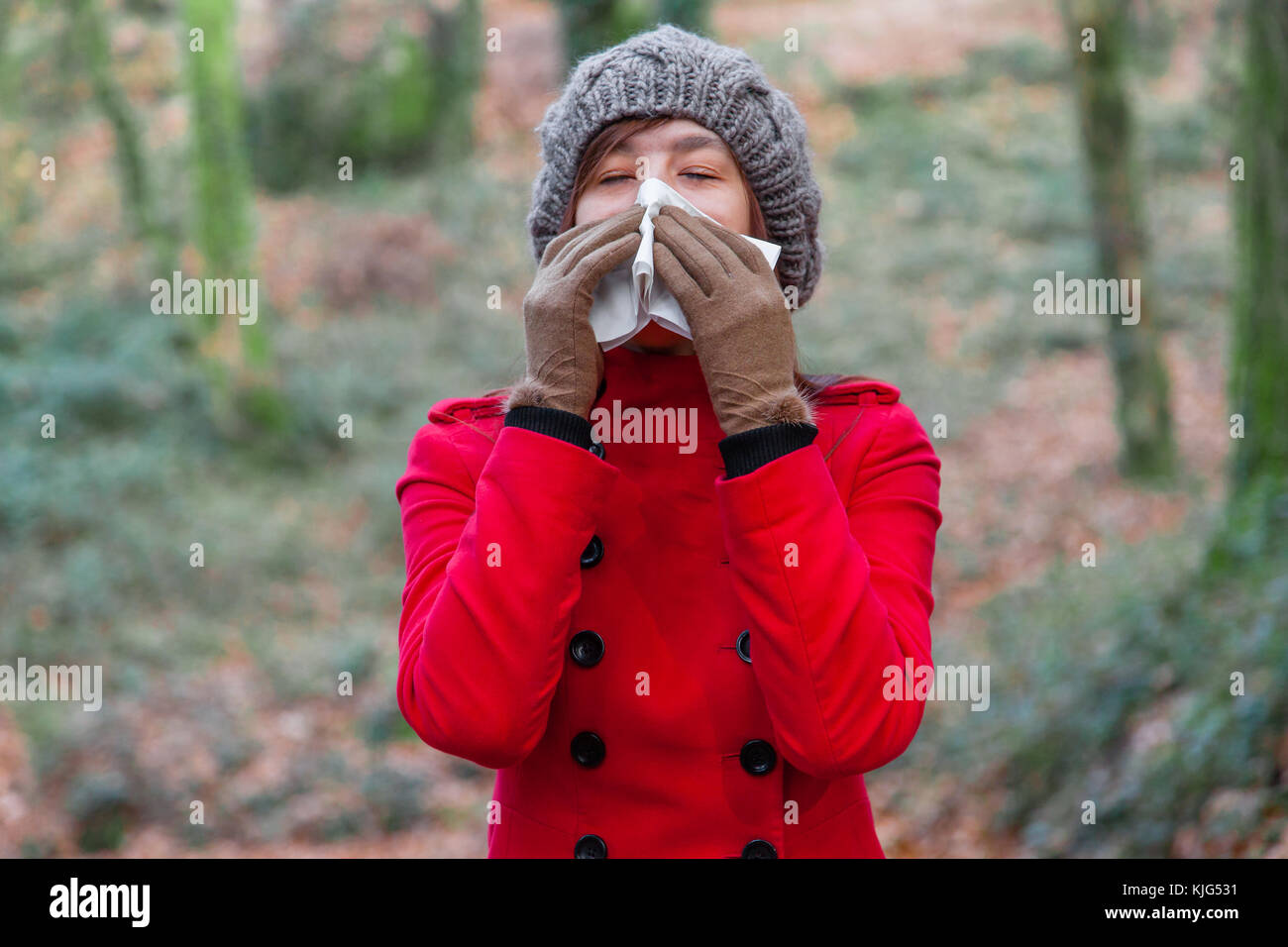 Junge Frau mit Erkältung oder Grippe blasen Nase, Niesen auf weißem Papier Taschentuch in Wald trägt einen roten Mantel oder Jacke und Mütze Stockfoto