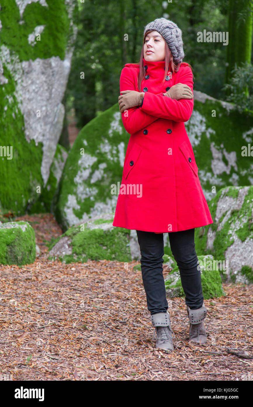 Junge Frau auf einem Wald zittern mit kalten und umarmen sich Holding verloren, trug einen roten Mantel oder Jacke, eine Mütze und Handschuhe während der Fal Stockfoto
