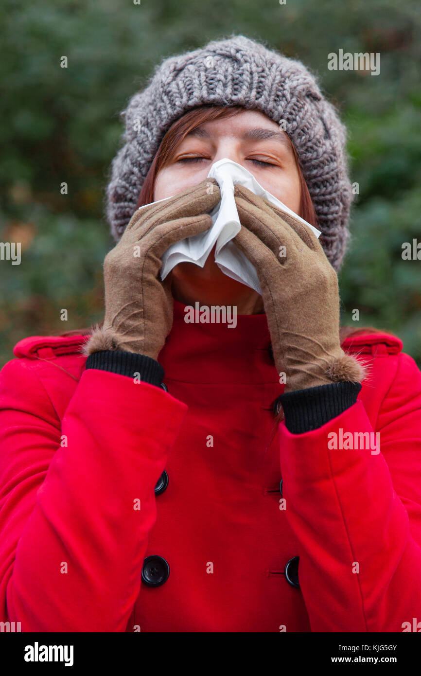 Junge Frau mit Erkältung oder Grippe blasen Nase, Niesen auf weißem Papier Taschentuch in Wald trägt einen roten Mantel oder Jacke, eine Mütze ein Stockfoto