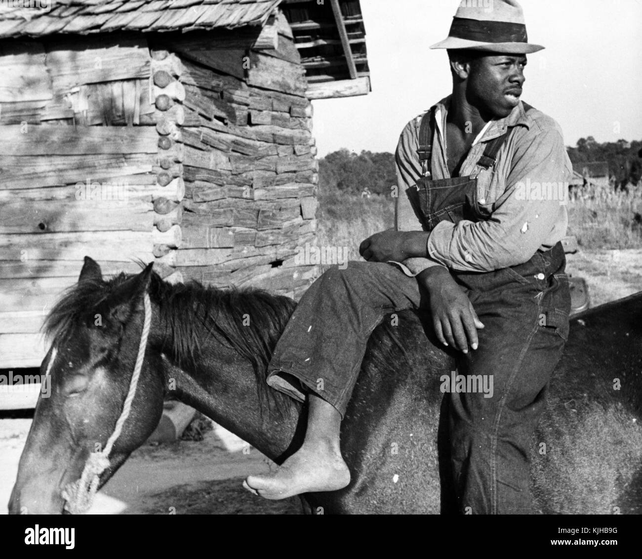 Ein Foto von einem Mann in Jeans Overalls gekleidet, ein Hemd und Hut sitzt auf dem Rücken eines Pferdes, er ist eine Erdbeere Picker, der seine Schuhe entfernt hat, während sie eine Pause, eine hölzerne Gebäude und Felder hinter ihnen gesehen werden können, Hammond, Louisiana, 1935. Von der New York Public Library. Stockfoto