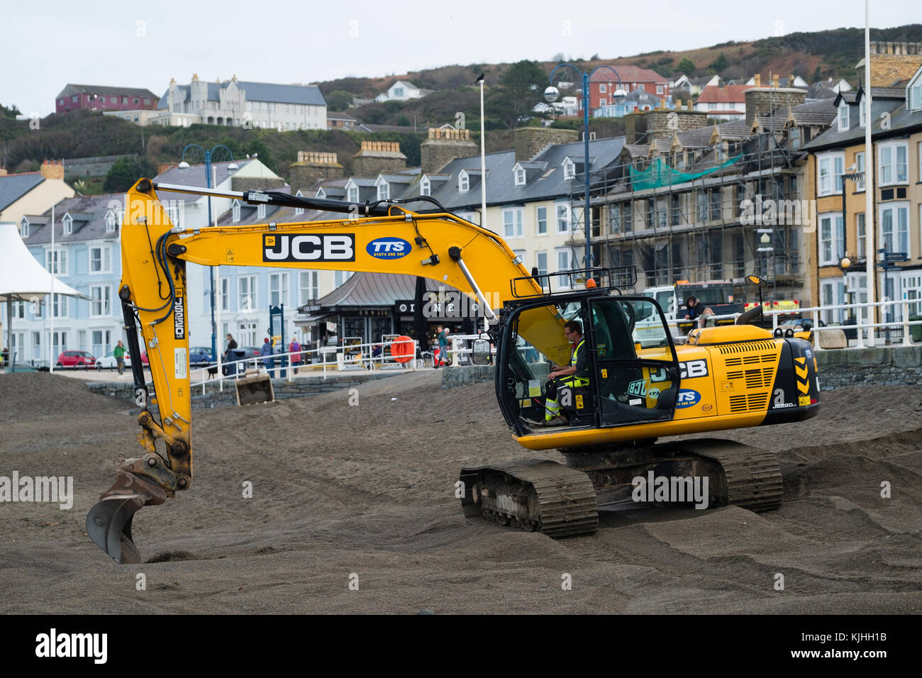 Nach dem Sturm: Ein JCB-Bagger von den Werkunternehmen TTS, mit lokalen Behörden, die Spaten verwenden, um Tonnen Sand von der Küste von Aberystwyth zu entfernen. Der Sand war während des Sturms Brian aufgespült worden und musste in der Höhe reduziert werden, um sicherzustellen, dass die Meeresmauer eine wirksame Barriere für die nächste Sturmflut und Flut blieb Stockfoto