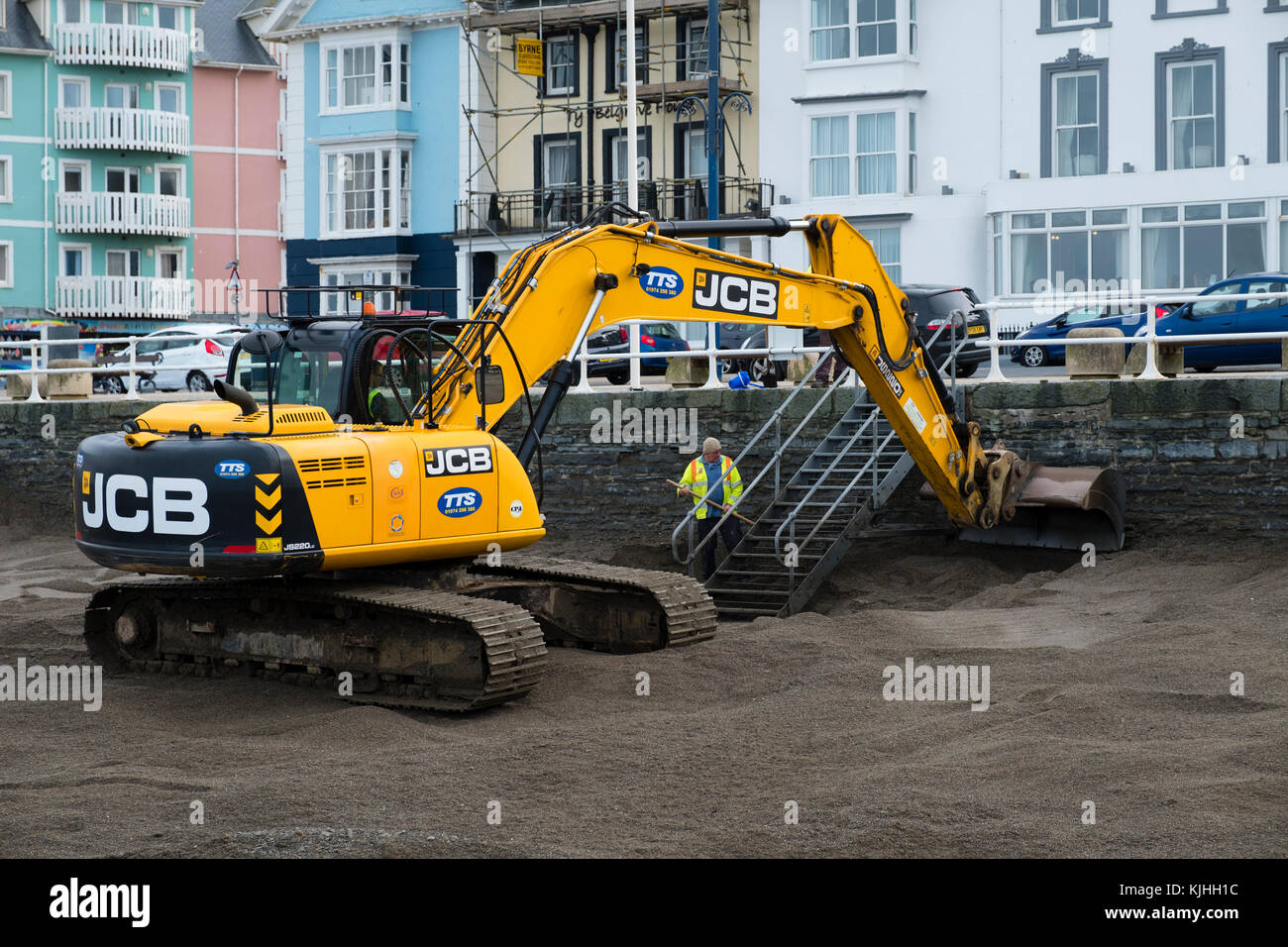 Nach dem Sturm: Ein JCB-Bagger von den Werkunternehmen TTS, mit lokalen Behörden, die Spaten verwenden, um Tonnen Sand von der Küste von Aberystwyth zu entfernen. Der Sand war während des Sturms Brian aufgespült worden und musste in der Höhe reduziert werden, um sicherzustellen, dass die Meeresmauer eine wirksame Barriere für die nächste Sturmflut und Flut blieb Stockfoto