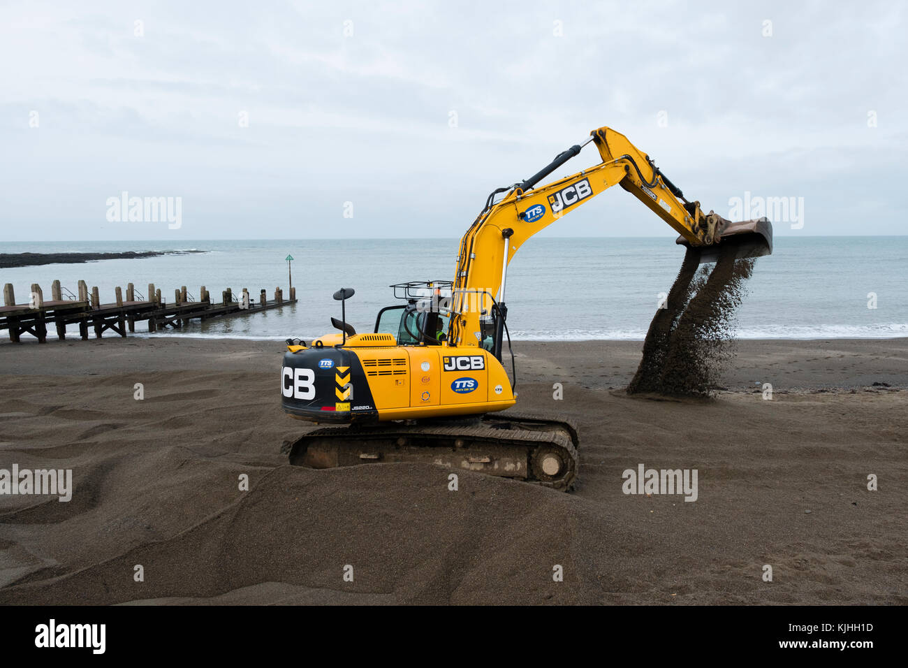 Nach dem Sturm: Ein JCB-Bagger von den Werkunternehmen TTS, mit lokalen Behörden, die Spaten verwenden, um Tonnen Sand von der Küste von Aberystwyth zu entfernen. Der Sand war während des Sturms Brian aufgespült worden und musste in der Höhe reduziert werden, um sicherzustellen, dass die Meeresmauer eine wirksame Barriere für die nächste Sturmflut und Flut blieb Stockfoto