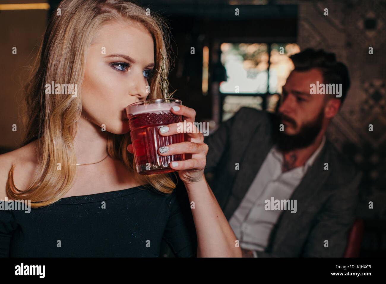 Frau trinkt einen Cocktail in einer Bar mit einem Mann hinter ihr sitzen Stockfoto