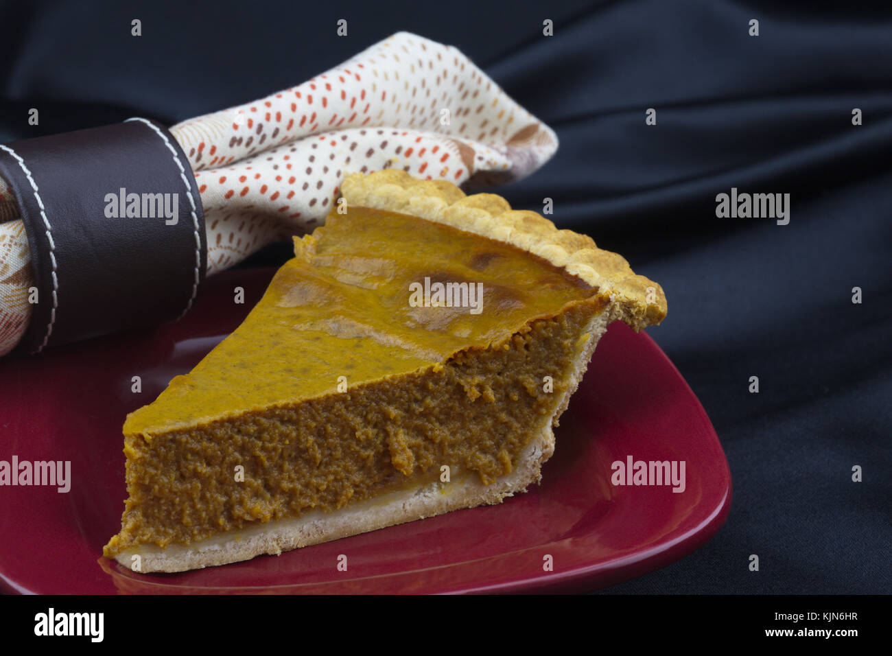Köstliches Stück feucht Pumpkin Pie mit zarten Gebäck Kruste auf rote Platte akzentuiert durch die Farben des Herbstes Serviette und braunem Leder ring gegen schwarze Sati Stockfoto