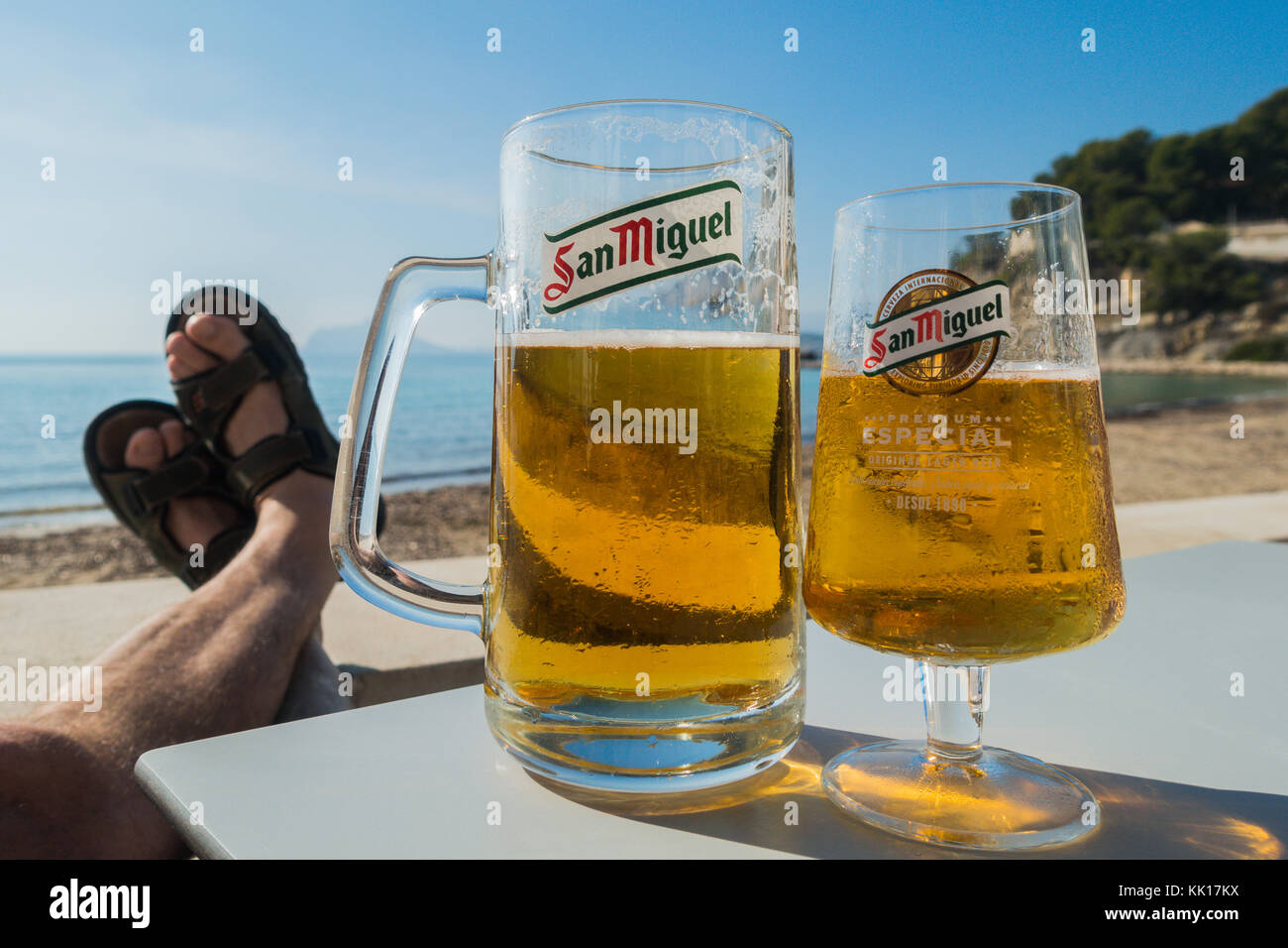 Zwei Gläser San Miguel Bier halb voll, ein großes und ein kleines, mit mans Füße in Sandalen aufgestützt am Meer in der Sonne Stockfoto