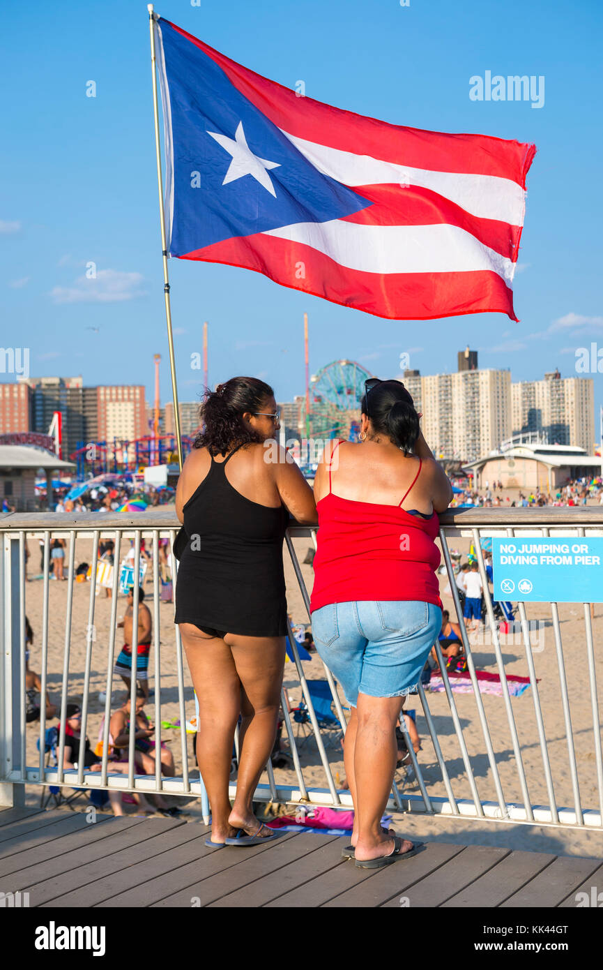 New York City - 20. August 2017: ein Puerto Rican flag Wellen über zwei Frauen, die auf der Promenade mit Blick auf den überfüllten Strand von Coney Island. Stockfoto