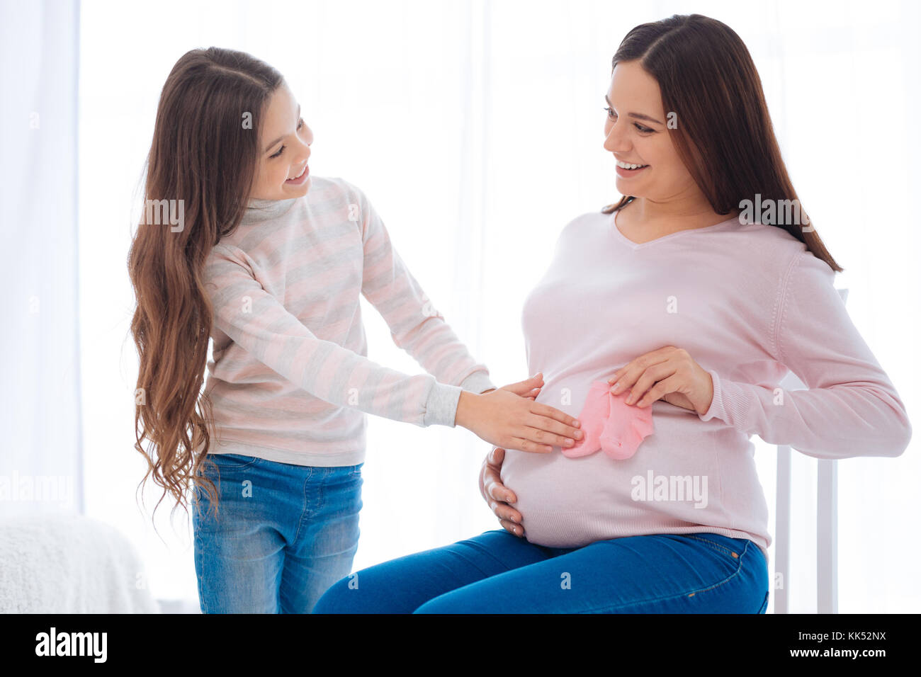 Pflege in alles. Angenehme glücklich strahlenden Mädchen schwangere Mütter Bauch streicheln, während sie Holding rosa Socken und auf dem Stuhl Stockfoto