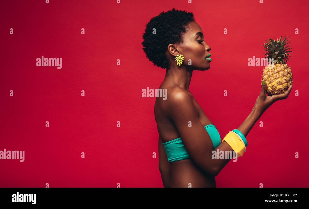 Seitenansicht der schwarzen Frau an Ananas in der Hand. Weibliche Mode Modell mit lebendigen Make-up über den roten Hintergrund. Stockfoto