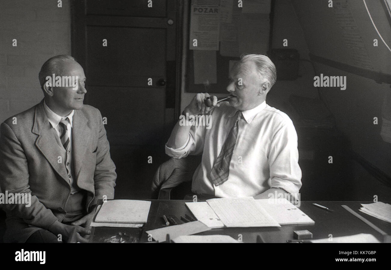 1950er Jahre, historische Bild, das einen Mann zeigt in seinem Hemd und Krawatte an seinem Schreibtisch in einem Büro Rauchen eine Tabakpfeife sitzt und in einer Diskussion mit einem anderen Mann. Pippe Rauchen wurde unter erwachsenen Männern in Großbritannien zu dieser Zeit populär. Stockfoto