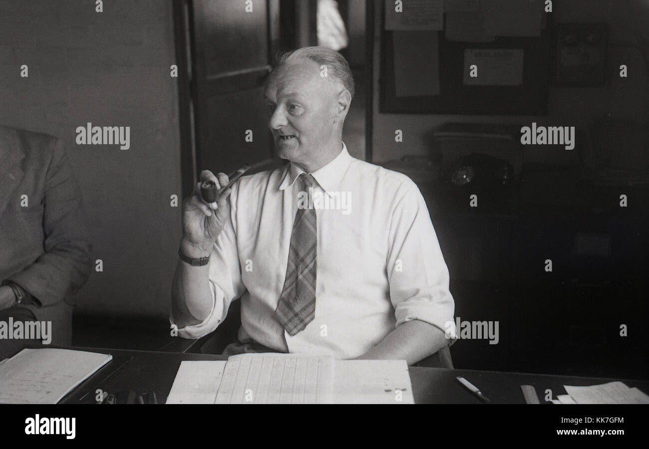 1950er Jahre, historische Bild, ein Mann am Schreibtisch in einem Büro mit einem Rohr in der Hand, England, Großbritannien sitzen. Mit einem Rohr ist eine traditionelle Form des Rauchens Tabak und war unter den Menschen einer bestimmten Altersgruppe zu dieser Zeit populär. Stockfoto