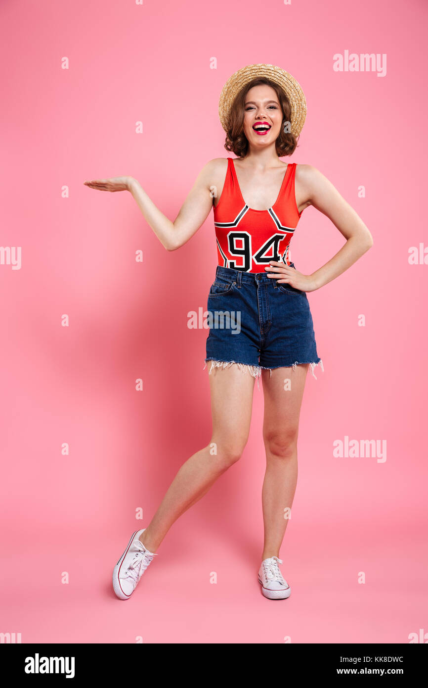 Volle lengrh Portrait von fröhlichen Irl in rot Bodys und Jeans kurze stehen mit Hand auf ihre Hüfte, Anzeigen leer Palm, auf rosa Hintergrund isoliert Stockfoto