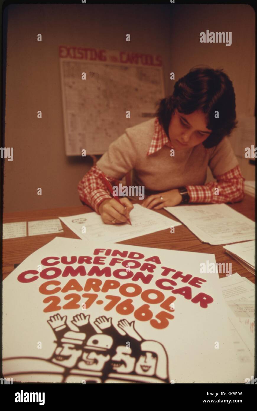 Farbfoto einer Frau ausfüllen Schreibarbeit am Schreibtisch, mit einem Schild mit der Aufschrift "Finde ein Heilmittel für die gemeinsamen Auto, Fahrgemeinschaft', 1974. Mit freundlicher Genehmigung der nationalen Archive. Stockfoto