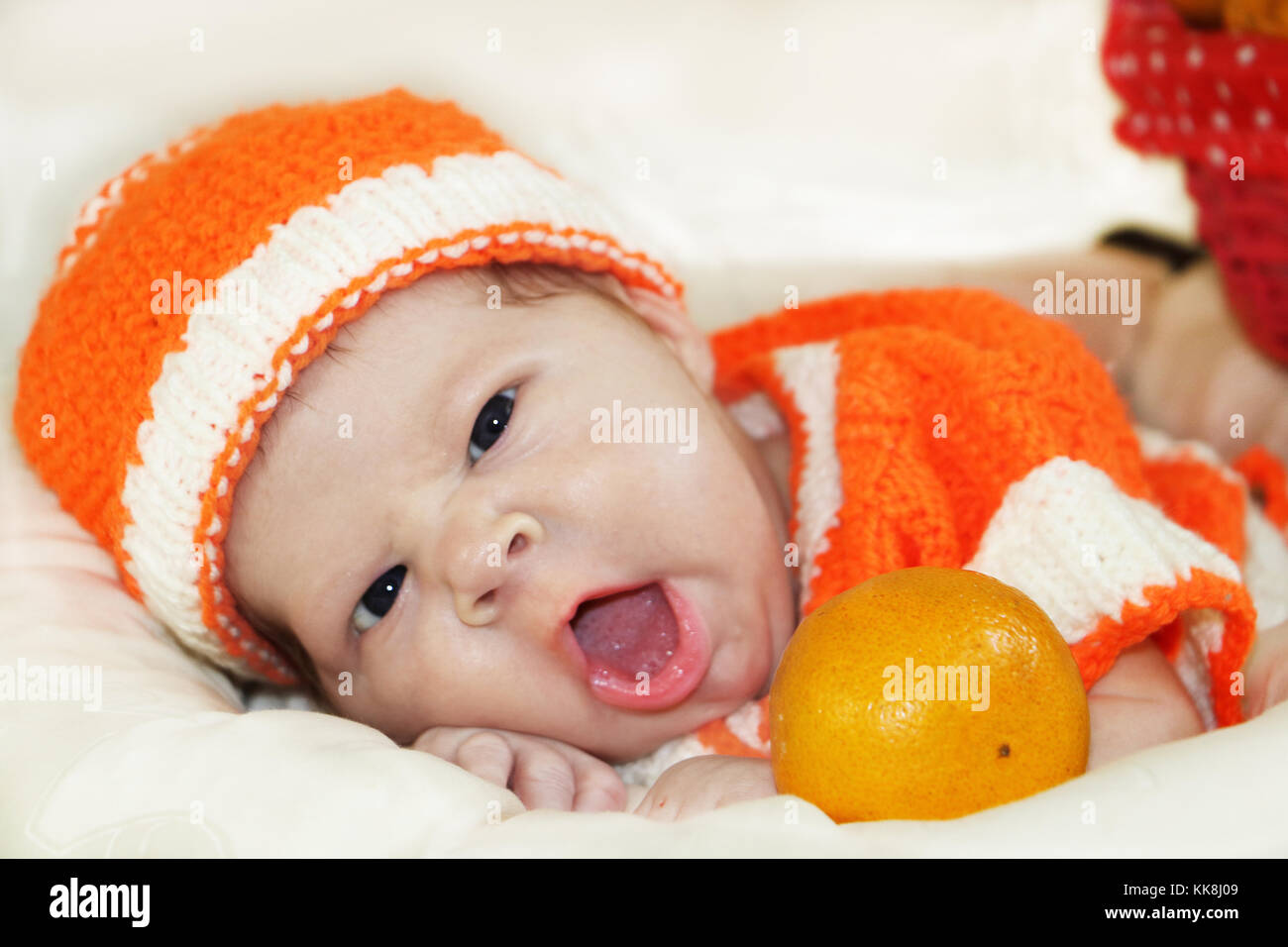 Cute Gähnen neugeborenes Baby in einen gestrickten orange Kostüm mit Orange in der vor ihm gekleidet. Herbst Halloween oder Ernte Konzept. Baby Gesicht expressi Stockfoto