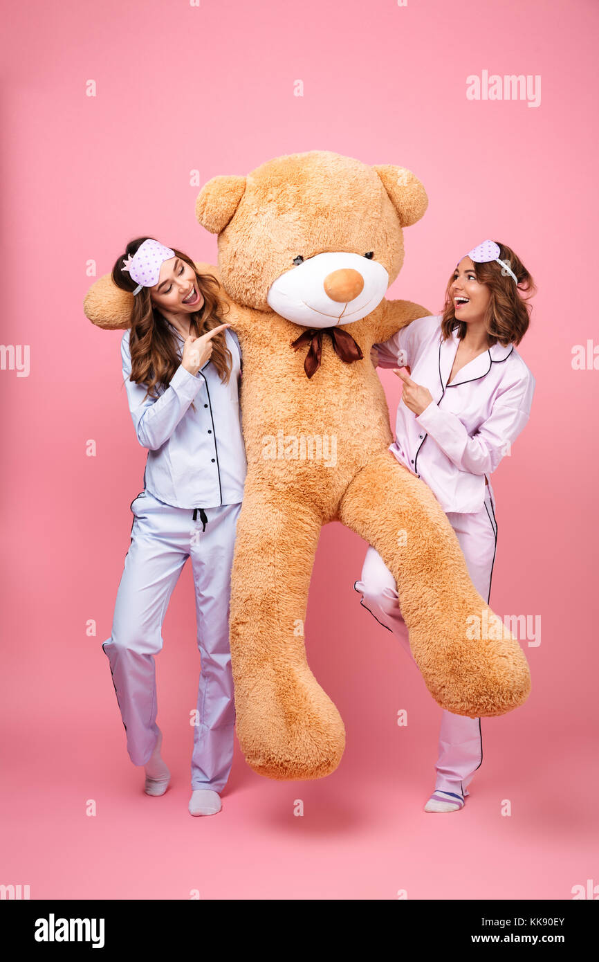 Foto von zwei erstaunliche Freunde Frauen im Schlafanzug über rosa Hintergrund hug Big teddy Spielzeug tragen isoliert. Neben der Suche zeigt. Stockfoto