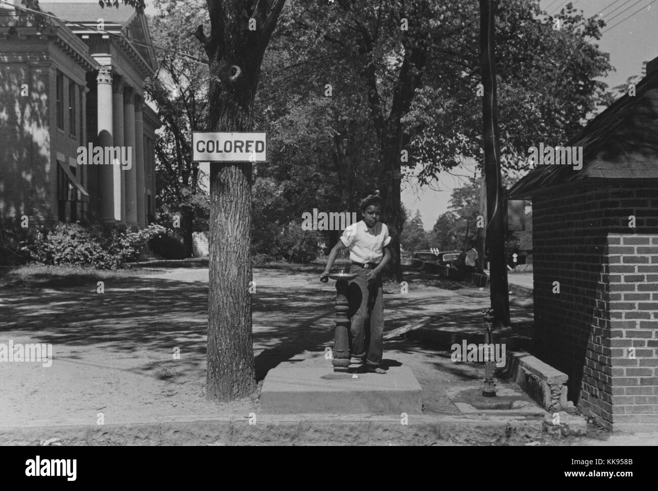 Ein Foto einer afrikanischen amerikanischen Kind mit einem Brunnen, der Baum neben dem Brunnen hat ein Schild angebracht, das liest "gefärbt", um Jim Crow Gesetze zu erzwingen, der Brunnen ist Teil des County courthouse Eigenschaft, Halifax, North Carolina, 1938. Von der New York Public Library. Stockfoto