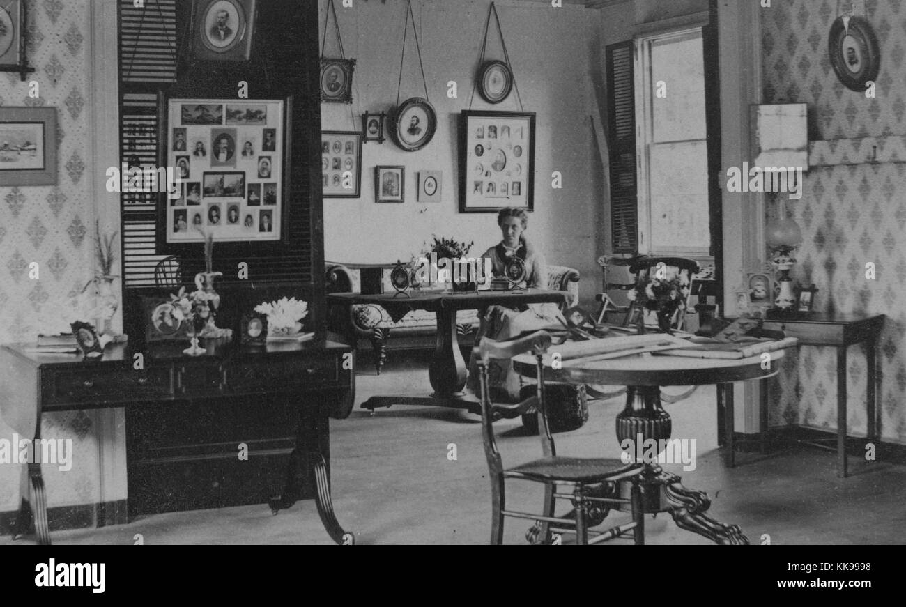Ein Foto von einer Frau, die an einem Tisch sitzen in einem Zimmer an der Rezeption, das Zimmer ist eingerichtet mit mehreren Frames, die Collagen von Fotografien enthalten, andere Bilder an den Wänden enthalten kleine Kunstwerke und Einzelporträts, Schreibtische und Tische in dem Zimmer dekorieren mit Fotografien, Blumen, Bücher und Schmuck, Antiqua, 1900. Von der New York Public Library. Stockfoto