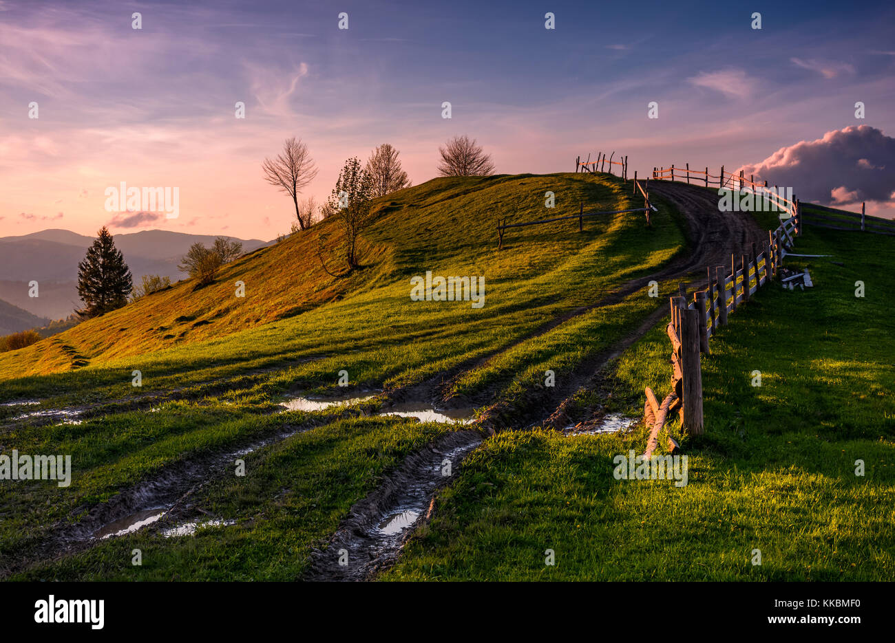 Holz Zaun entlang des Landes Schmutz Weg bergauf die grasbewachsenen Hügel im Frühling in der Abenddämmerung. spektakuläre Natur Landschaft in bergigen ländlichen Gegend mit Gorg Stockfoto