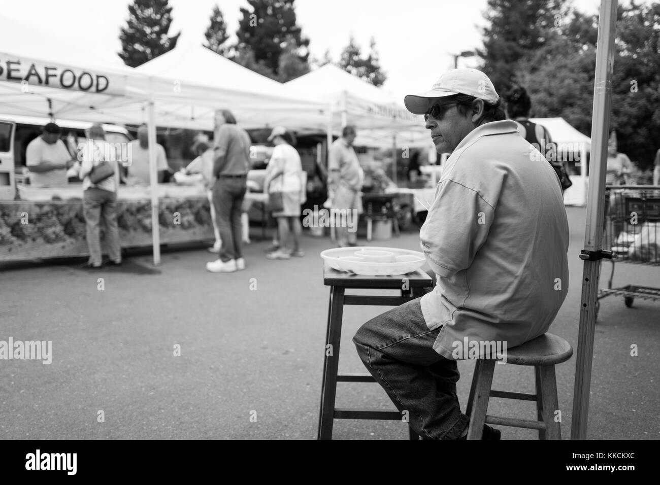 Auf einem wöchentlichen Bauernmarkt in der San Francisco Bay Area sitzt ein Händler mit Baseballhut und Sonnenbrille auf einem Hocker vor seinem Stand und bereitet sich darauf vor, an vorbeifahrende Kunden Fruchtproben zu verteilen, während die Leute an anderen Ständen im Hintergrund in Danville, Kalifornien, einkaufen. Juni 2016. Stockfoto