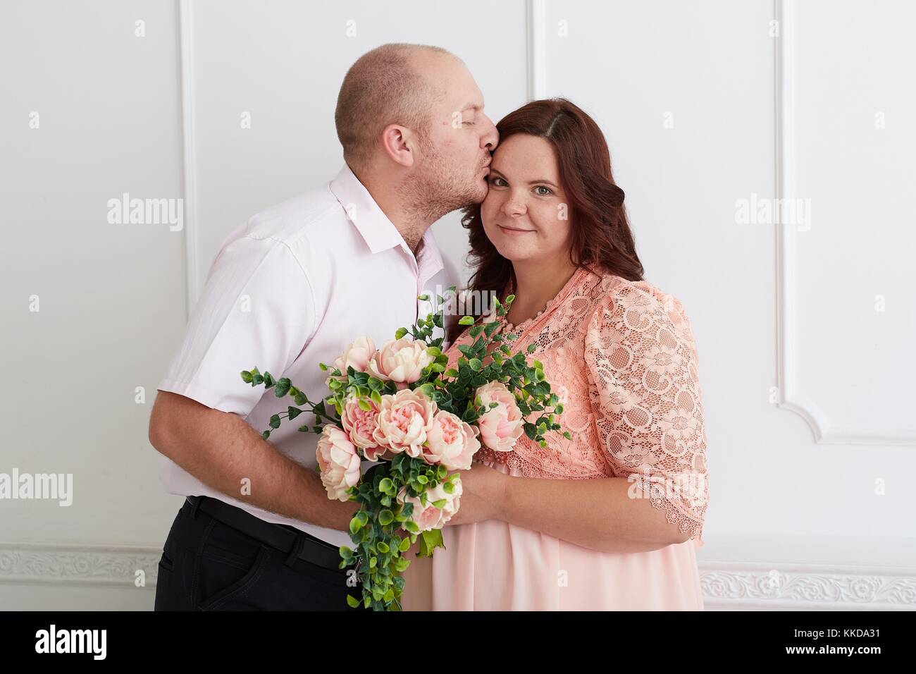 Mann küssen Lächeln schwangere Frau in Coral Kleid, die Bouquet Rose und Pfingstrose. Gerne küssen paar auf Luxus weiße Wand. Stockfoto