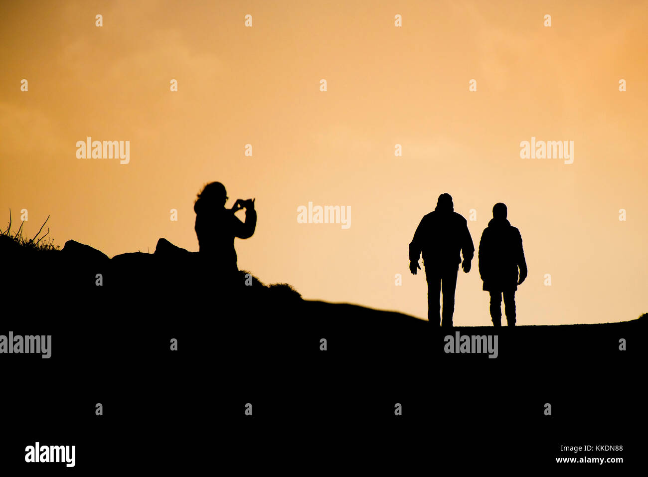 Silhouetten - eine Frau, die ihr Smartphone benutzt, um ein Paar zu fotografieren, das im späten Abendlicht läuft. Stockfoto