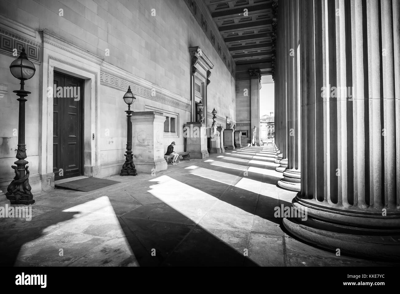 Licht und Schatten spielen auf der Säulen und die Architektur von St. George's Hall in lverpool Lime Street, North West England, mit einem Mann auf der integrierten Bank am oberen Ende der Stufen sitzend, auf einem schönen Sommer sonnigen Tag.. Stockfoto