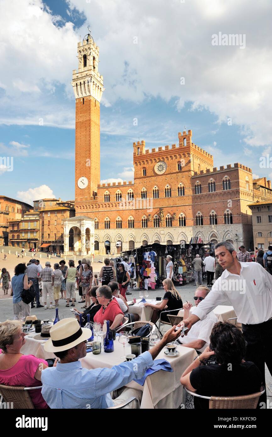 Touristen in Café am Piazza del Campo, dem zentralen Platz der Stadt Siena, Toskana, Italien. Torre del Mangia Turm erhebt sich hinter Stockfoto