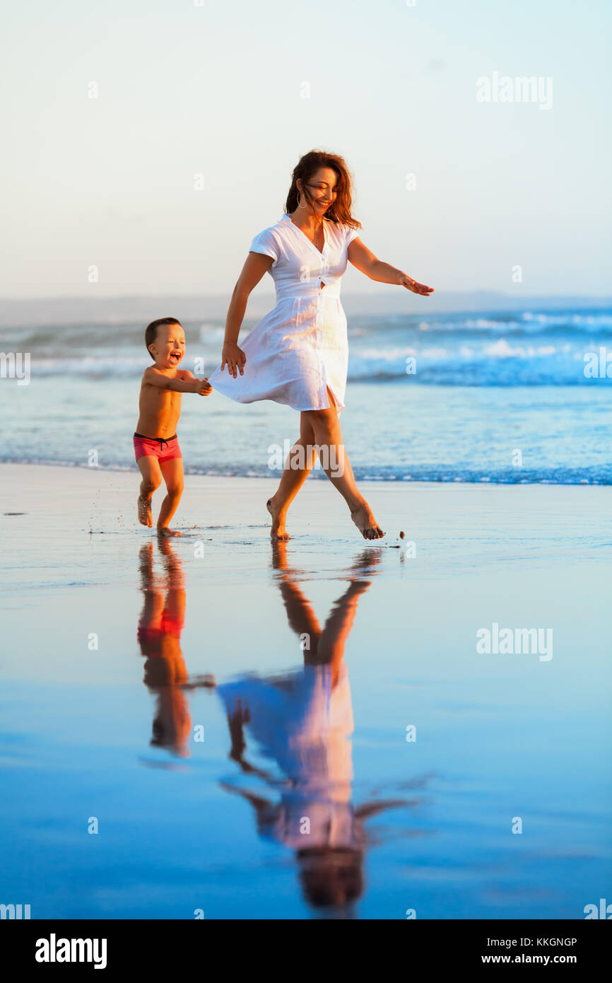 Glücklicher Familienurlaub - Mutter, Baby, Sohn, haben Spaß zusammen, barfuß, Kind wird am Wasserpool entlang der Sonnenuntergangswelle am Sandstrand geführt. Reise-Lifestyle Stockfoto
