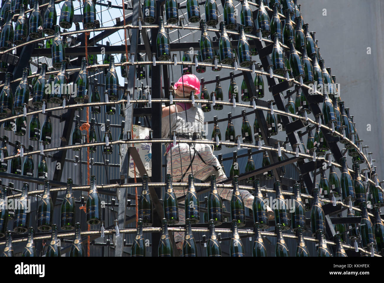 Kapstadt, Südafrika. 1.. Dezember 2017. Eine Frau mit Sicherheitsgeschirr und pinkem Hut, die an der V&A Waterfront in Kapstadt, Südafrika, einen Weihnachtsbaum aus beleuchteten Moet Chandon Champagnerflaschen baut. Stockfoto