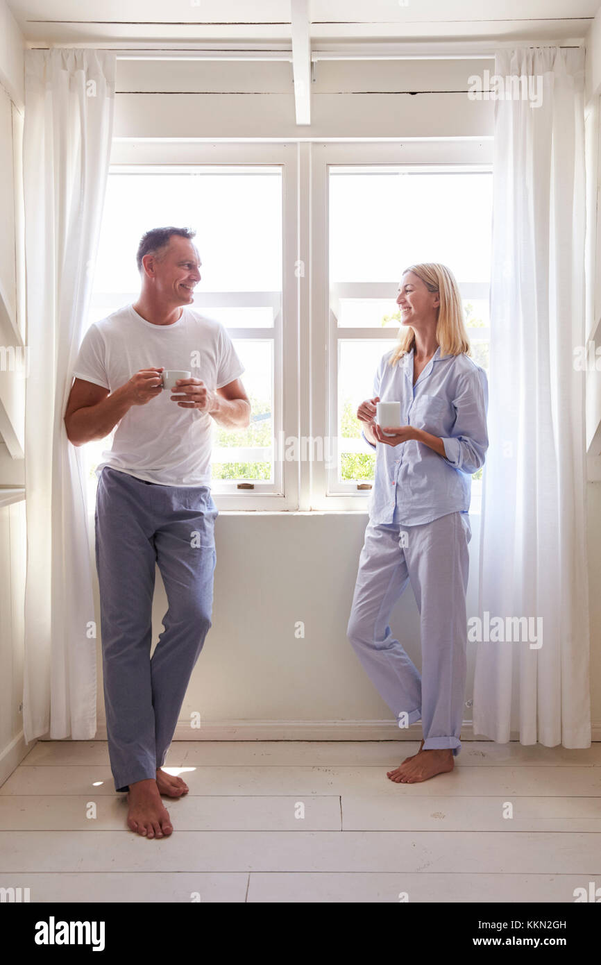 Reifes Paar stehend durch Schlafzimmer Fenster mit heißen Getränken Stockfoto
