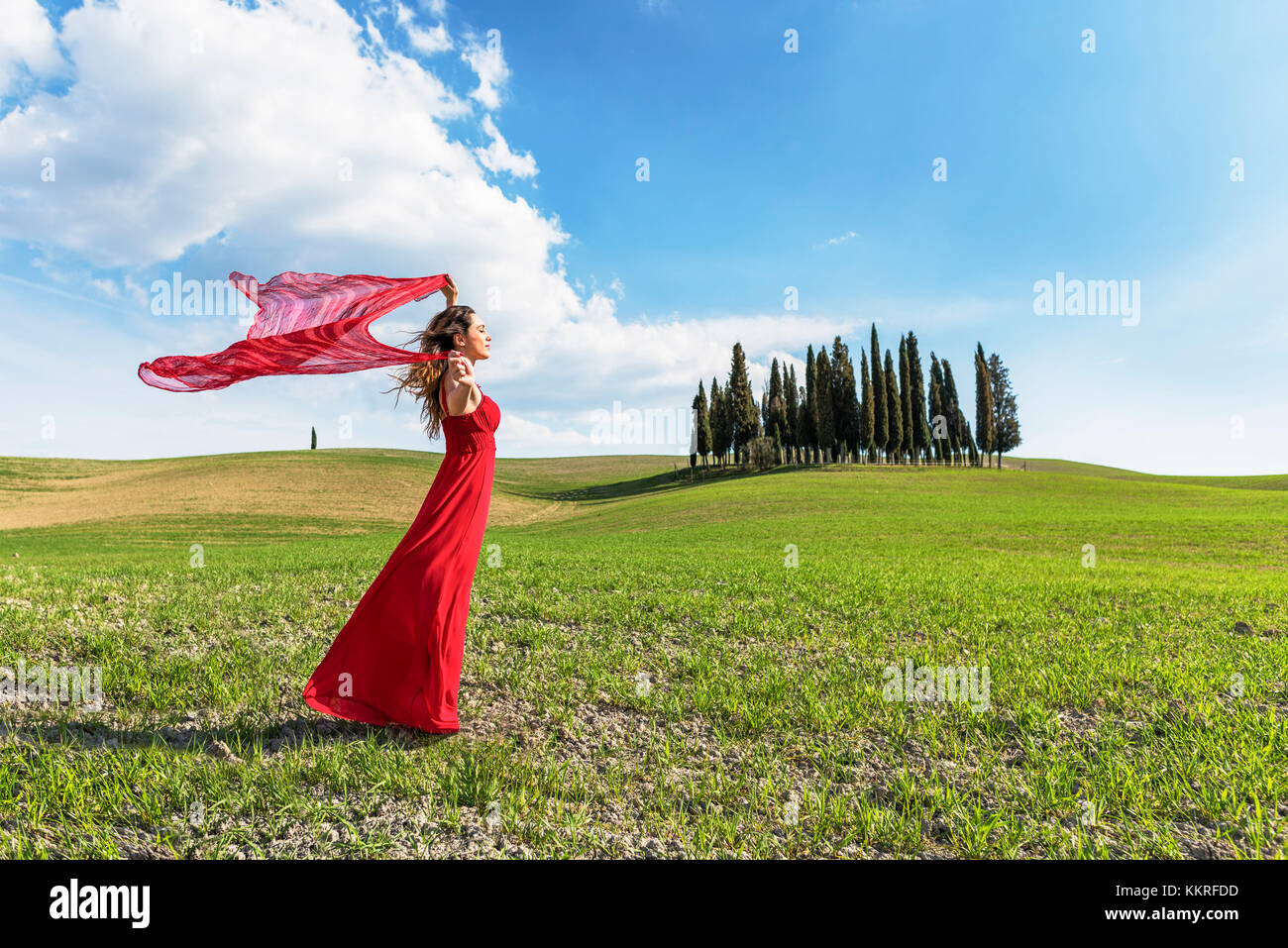 San Quirico d'Orcia, Val d'Orcia, Siena, Toskana, Italien. Eine junge Frau im roten Kleid entspannen in einem Weizenfeld in der Nähe der Zypressen des Orcia Tal Stockfoto