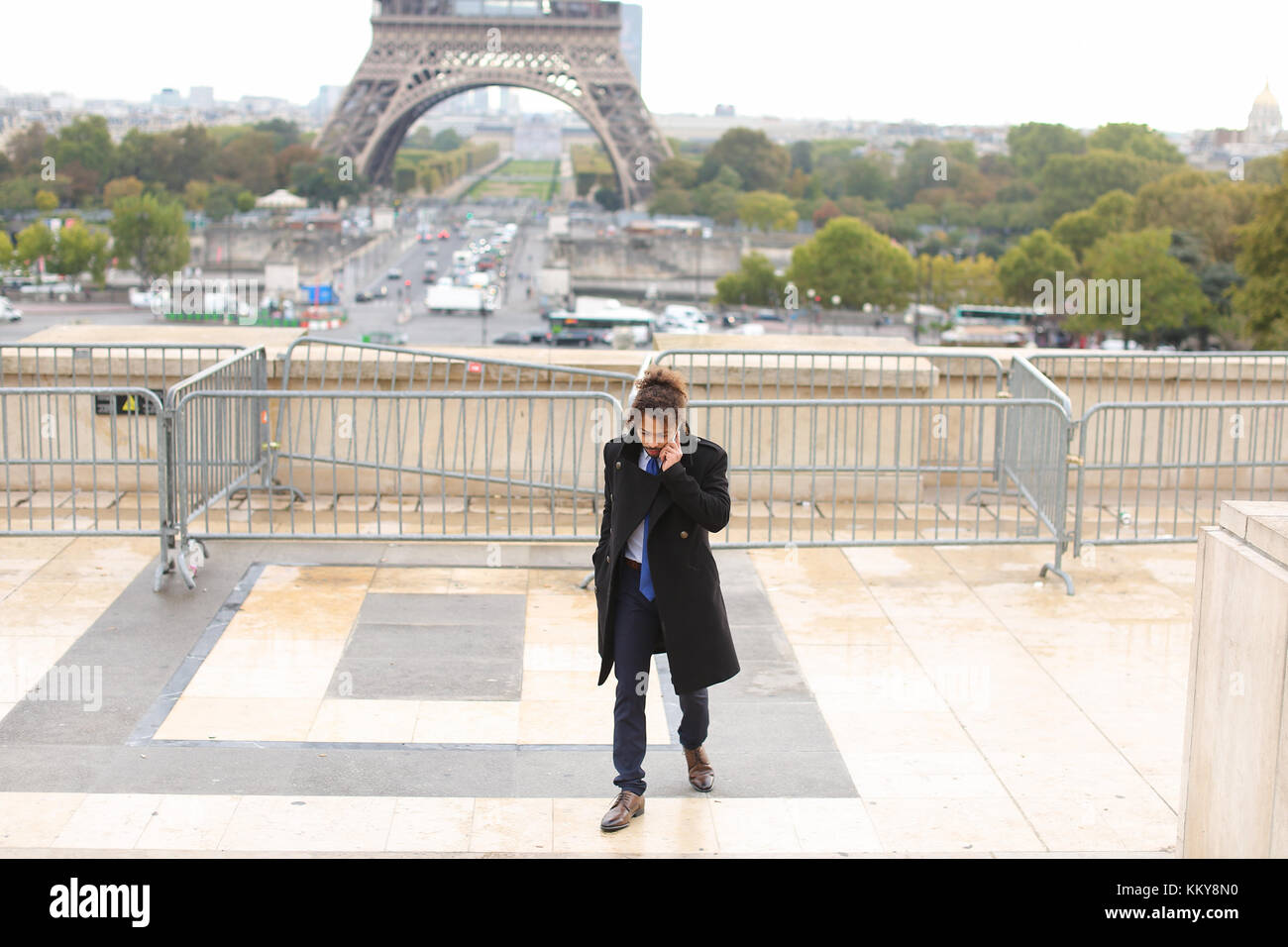 Brasilianische boy Chatten mit europäischen Freunden in der Nähe von Eiffelturm auf dem Smartphone. junger Mann im schwarzen Mantel bekleidet, hat earing, schwarzes lockiges Haar und Bart. Stockfoto