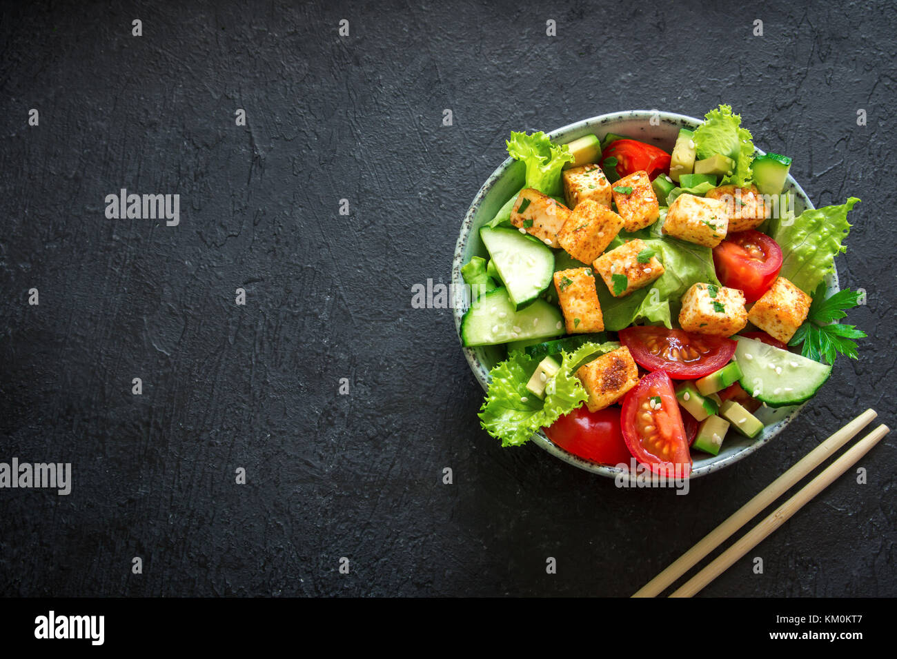 Gebratenem tofu Salat mit Gurken, Tomaten, Avocado und Sesamsamen. Hausgemachte asiatische Gemüse und Tofu Salat in Keramik Schüssel auf schwarzen Stein Hintergrund Stockfoto