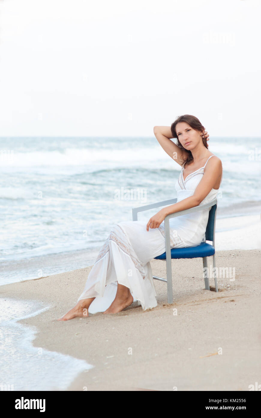 Junge Frau in weißem Kleid sitzt auf einem Stuhl am Strand, am Meer. romantische Bild Stockfoto