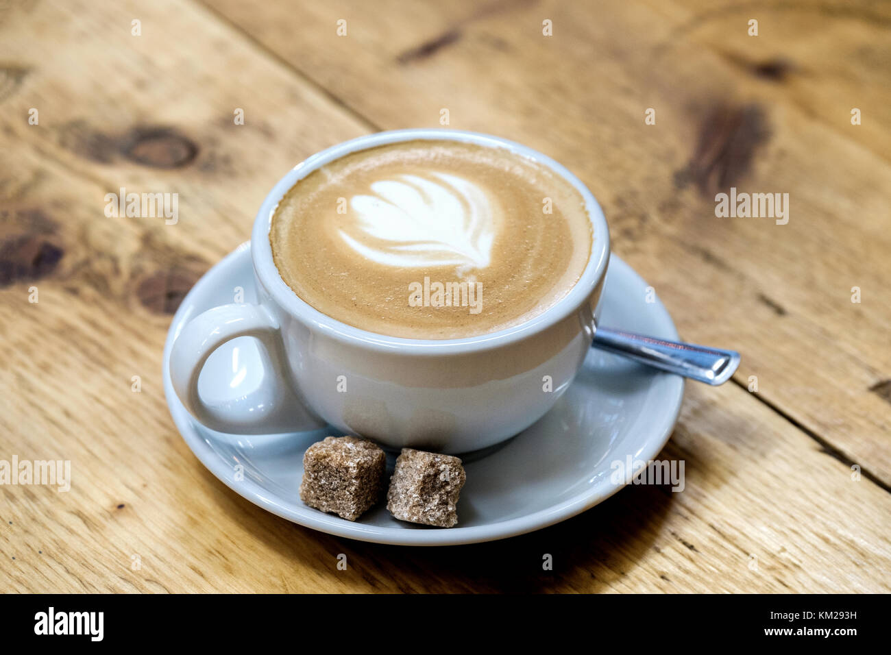 Ein frisch gebrauten Latte Kaffee in eine Tasse und Untertasse mit zwei Klumpen von braunem Zucker. Auf einem Tisch die Milch Kunstwerk in Form einer Tulpe gebildet wird gesetzt Stockfoto