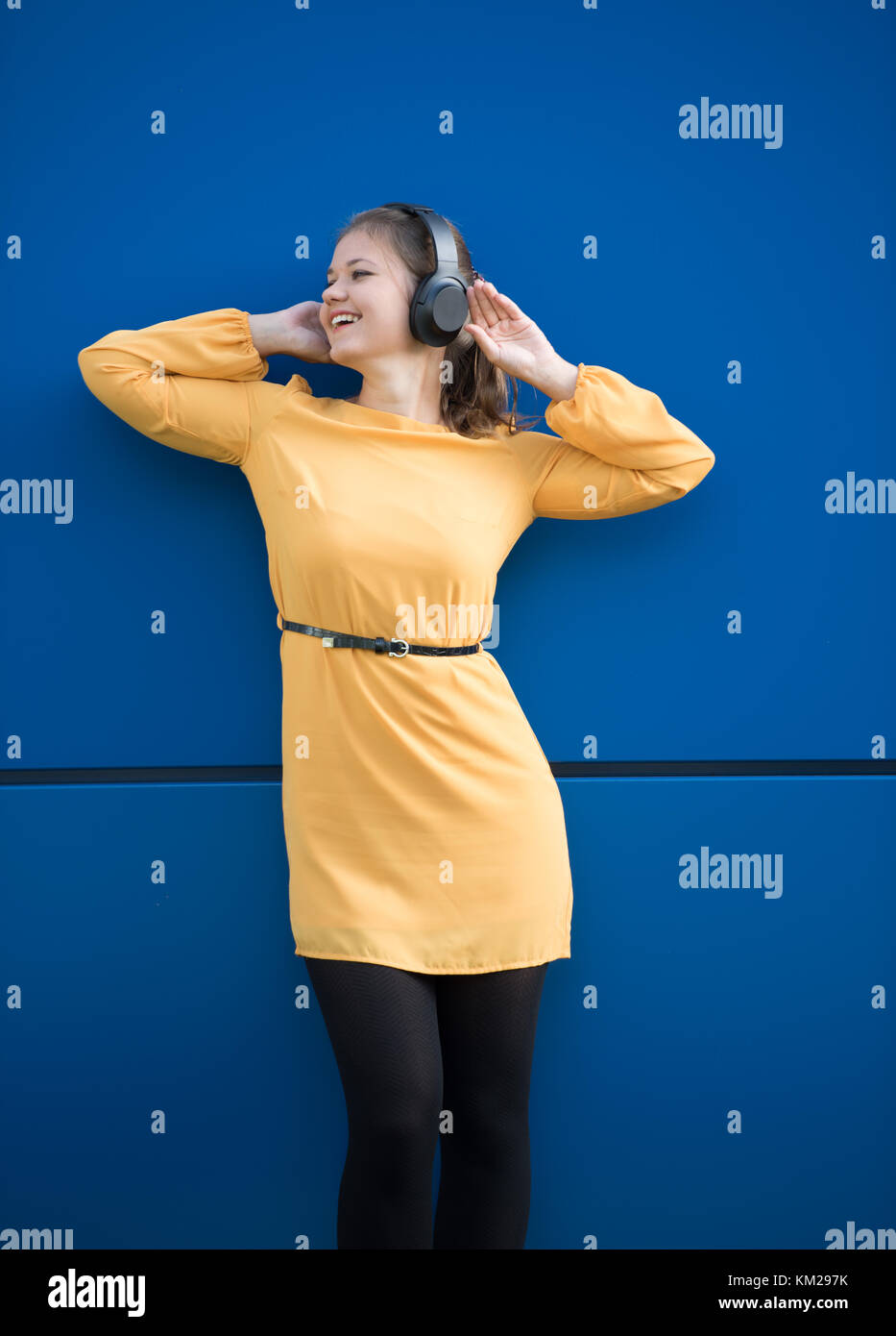 Junge hübsche Frau tragen gelbe Kleid Musikhören mit Kopfhörern vor blauem Hintergrund Stockfoto