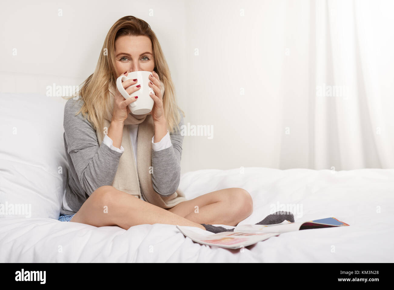 Attraktive blonde Frau mit einem reizenden Lächeln genießen Sie eine heiße Tasse Kaffee zu Hause an einem kalten Wintertag in ihrem warmen Trikot und Schal Stockfoto