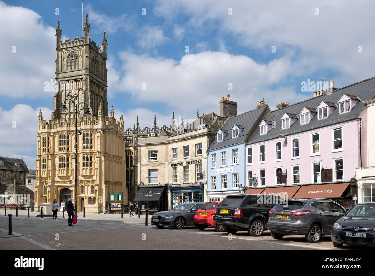 Den Marktplatz im Zentrum der Stadt Cirencester Gloucestershire, Vereinigtes Königreich. an einem sonnigen Tag, die Kirche des Hl. Johannes des Täufers. & Autos geparkt. Stockfoto