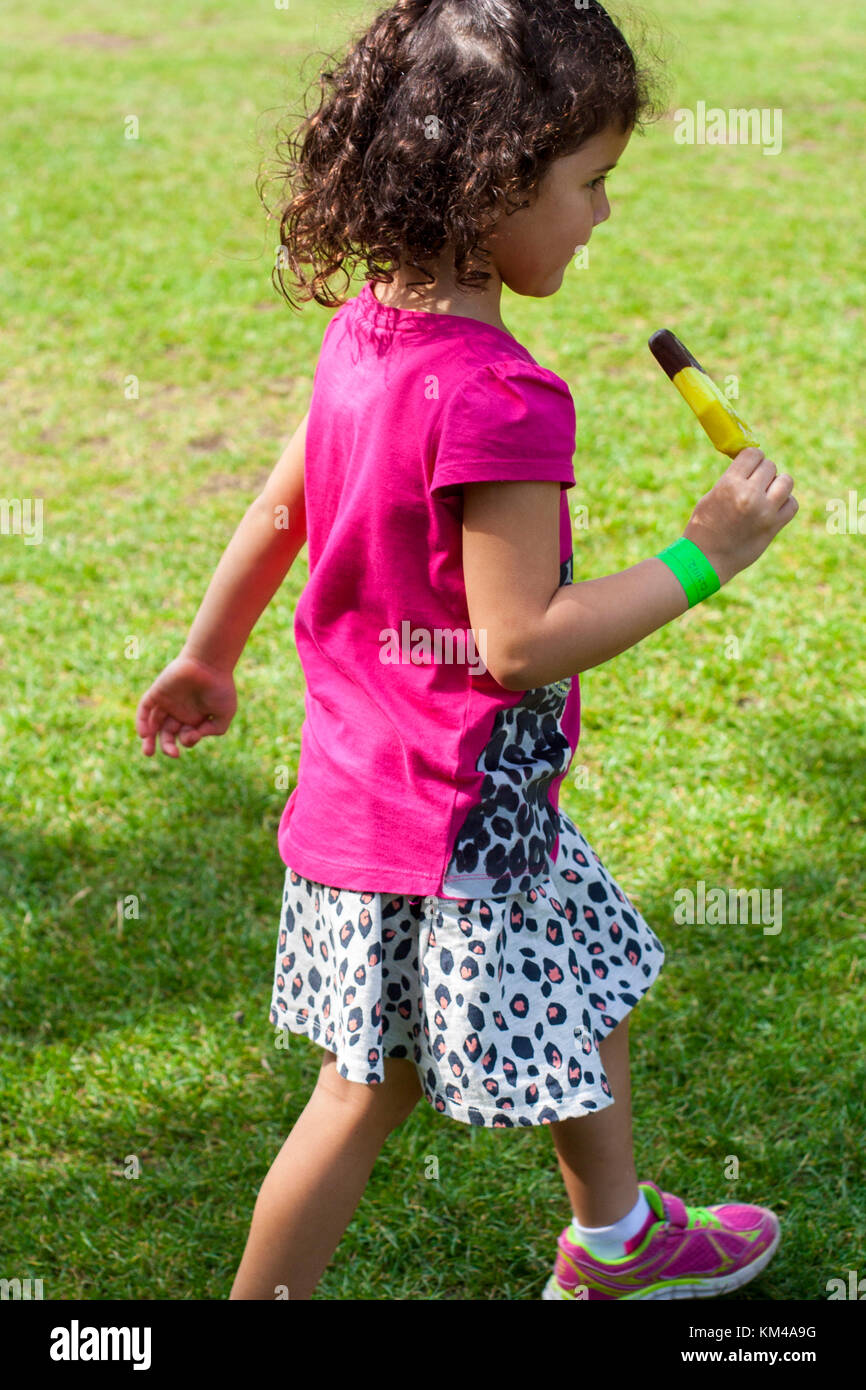 Kleines Mädchen mit schwarzen lockigen Haar Fuß in den Park Holding eine Schleife de Loop ice Pop im Sommer in Irland Stockfoto