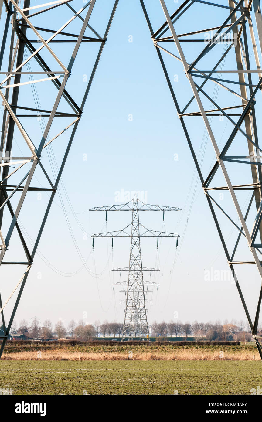 Eine Reihe von Hochspannung Strommasten stehen in einem ländlichen Gebiet und vor blauem Himmel in den Niederlanden. Stockfoto