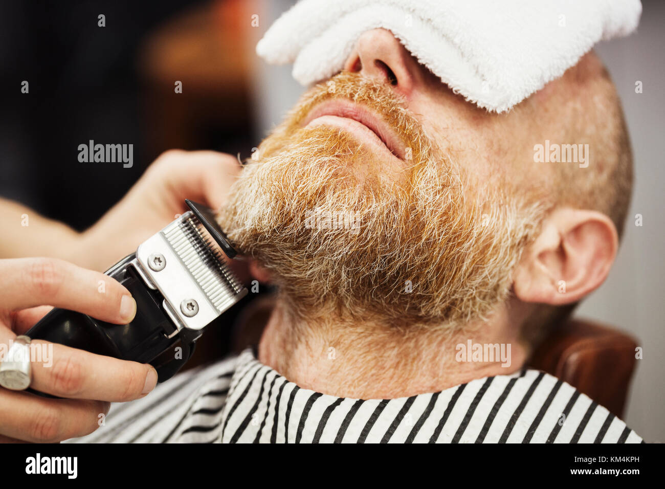 Ein Kunde in der Friseur Stuhl sitzen, mit einem warmen Handtuch auf sein Gesicht, und ein Friseur schneiden seinen Bart mit einem elektrischen Trimmer. Stockfoto