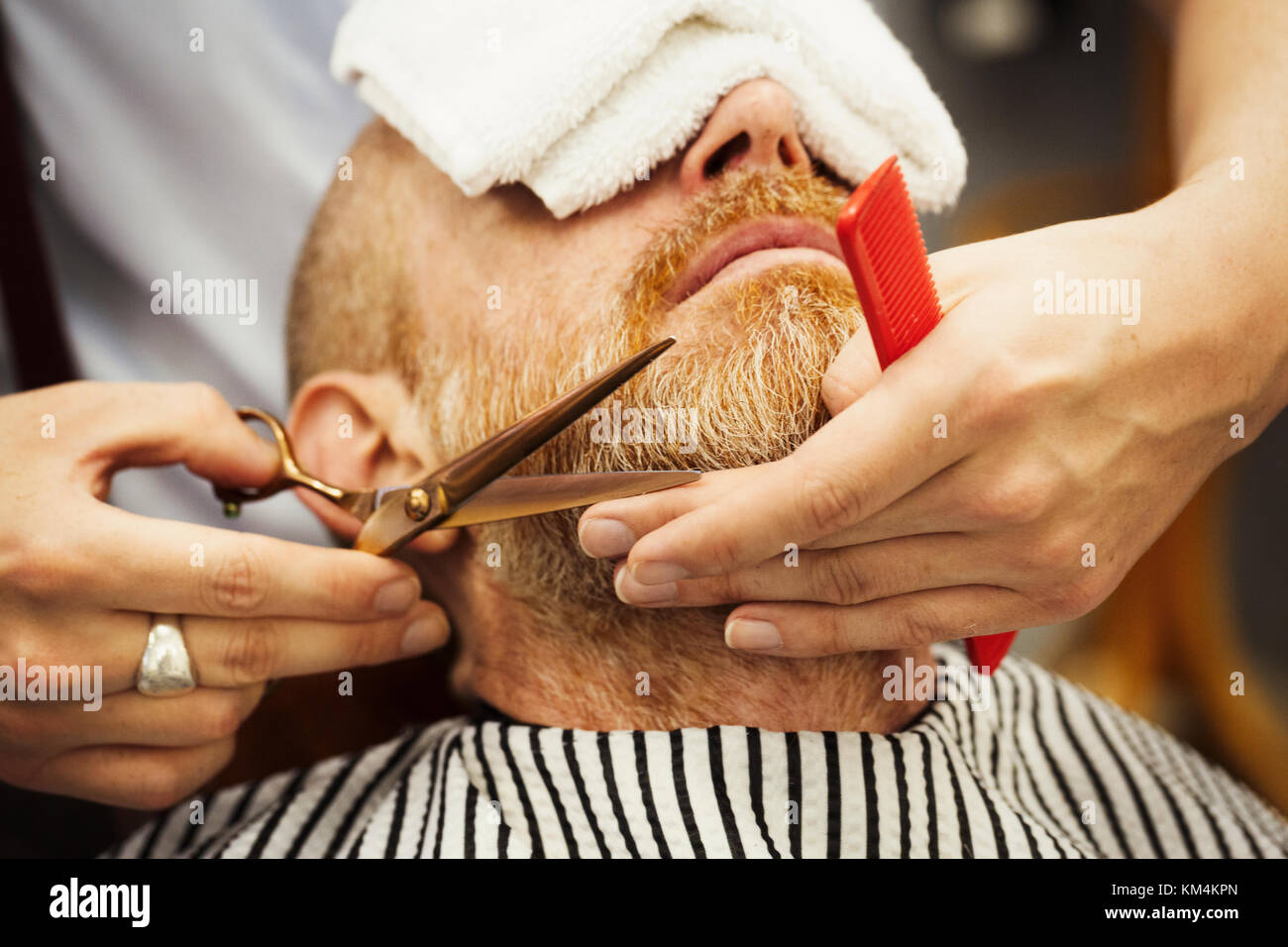 Ein Kunde in der Friseur Stuhl sitzen, mit einem warmen Handtuch auf sein Gesicht, und ein Friseur Schneiden von Bart. Stockfoto