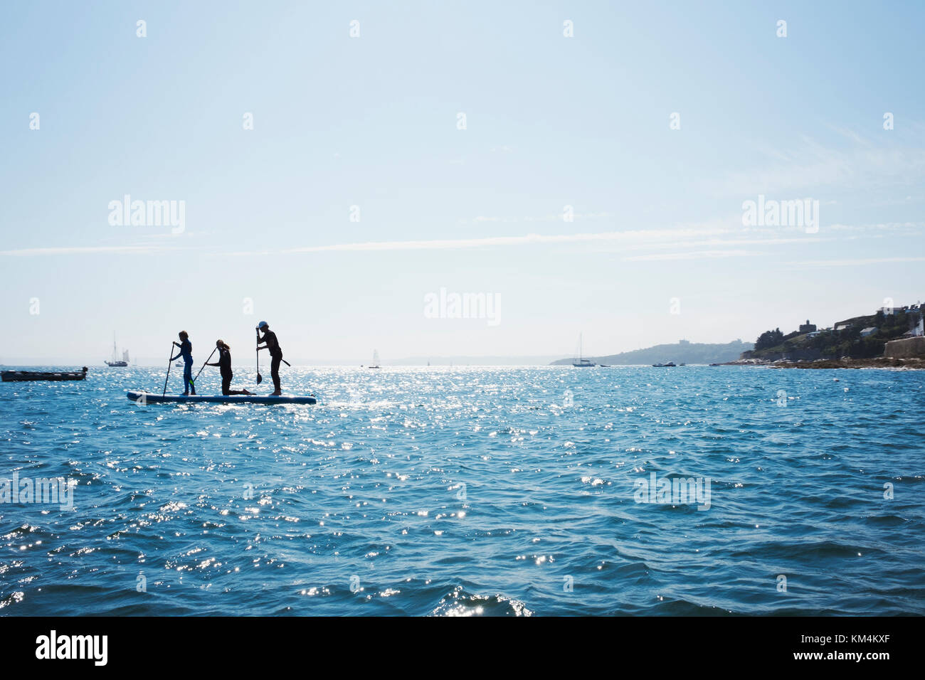 Silhouette von drei Menschen auf paddleboards auf dem Wasser, Aussicht auf das Meer von einem geschützten Abdeckung Yachten und Häuser auf der Landspitze zu günstig. Stockfoto