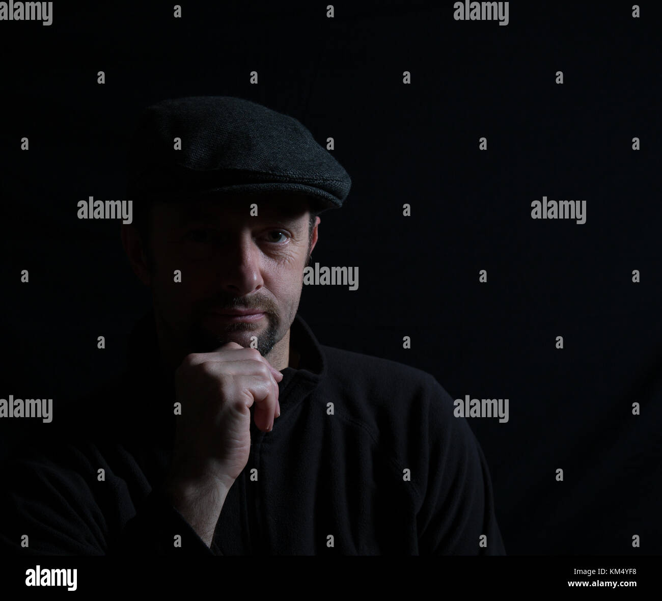 Künstlerisches Porträt eines isolierten Mannes in dunkler Mütze, halb beleuchtetes Gesicht, starrend auf Kamera, Hand auf Kinn, im Rampenlicht. Subtile Beleuchtung erzeugt ein Schattenbild. Stockfoto
